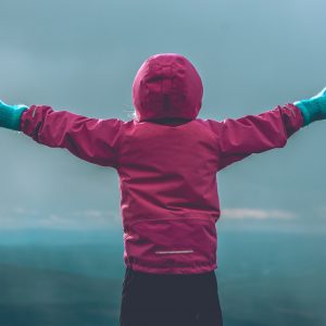 person wearing pink hooded jacket raising her hand in front of green mountain range during daytime