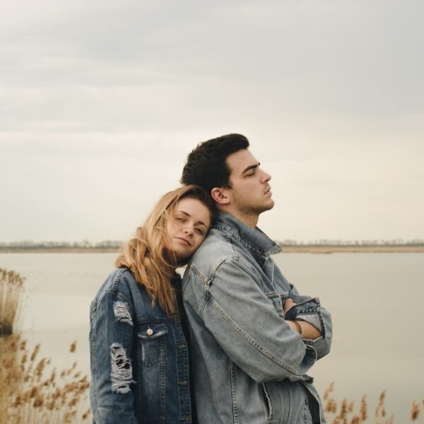 woman in blue denim jacket leaning on man's shoulder near body of water