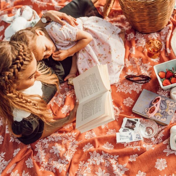 woman reading with girl while lying on orange and white floral picnic mat